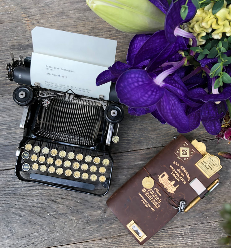 Corona 3 typewriter on wooden table. To right is a leatherbound traveler's journal. above right is a vivid purple orchid.